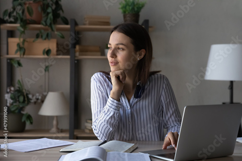 Fotografie Thoughtful millennial student girl sitting at table with laptop, open books, looking away, thinking over training course, learning webinar, online lesson, test