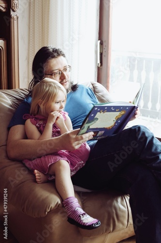 Dad reading bedtime story to his little daughter. They are sitting on big comfy armchair near window