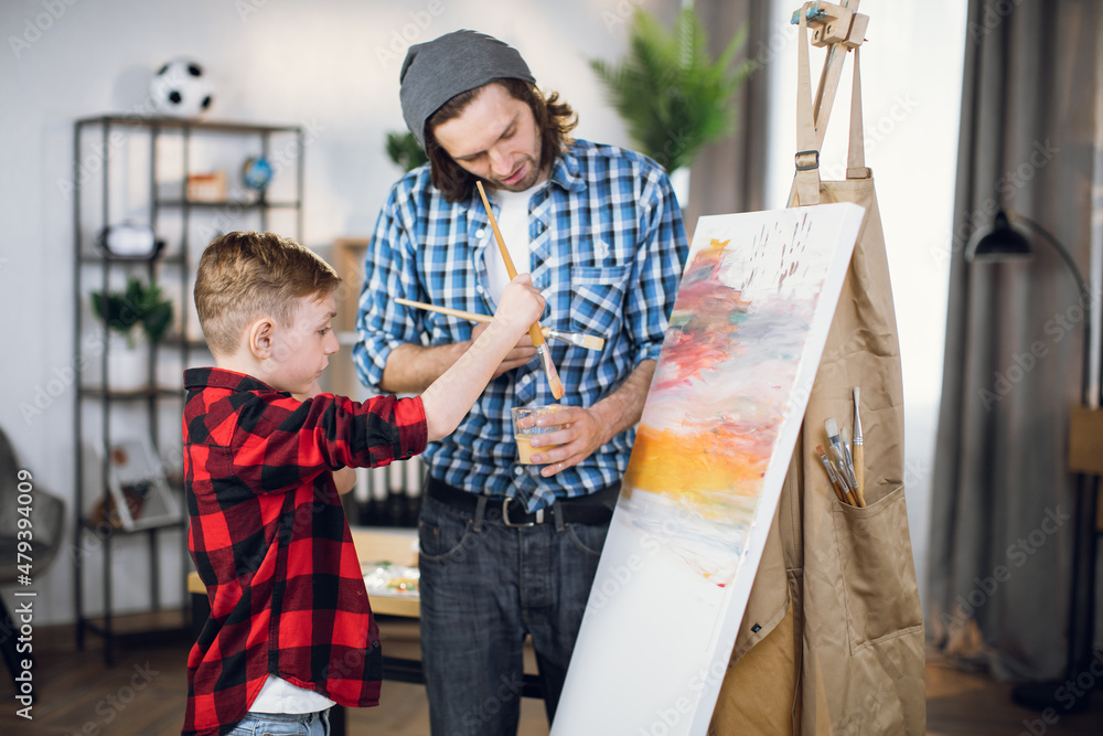 Handsome man and little boy in checkered shirts drawing abstract pattern on canvas. Happy father and son using brush and colorful paint during art process.