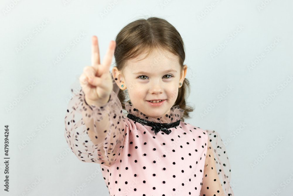Smiling little girl with two ponytails in a dress with polka dots on a white background shows two fingers raising her hand forward. Studio photo Smiling little girl with two ponytails in a dress with polka dots on a white background shows two fingers raising her hand forward. Studio photo