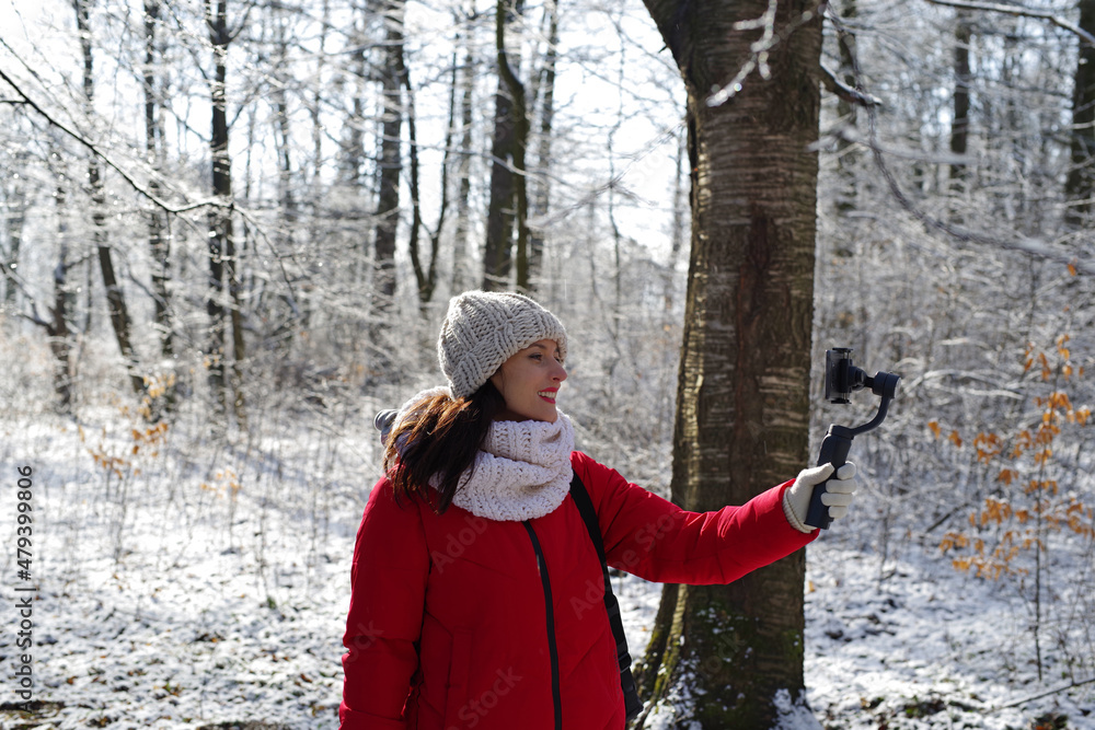 Woman blogger holding steadicam, smartphone, streaming in the woods in winter outdoors. Girl in red clothes and white hat hiking in snowy forest. Frosty sunny day. Female lifestyle portrait Woman blogger holding steadicam, smartphone, streaming in the woods in winter outdoors. Girl in red clothes and white hat hiking in snowy forest. Frosty sunny day. Female lifestyle portrait