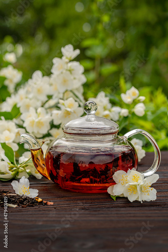 Steamed glass teapot with jasmine tea among flowering bushes. Outdoor  picnic  brunch. Floral background in blur.