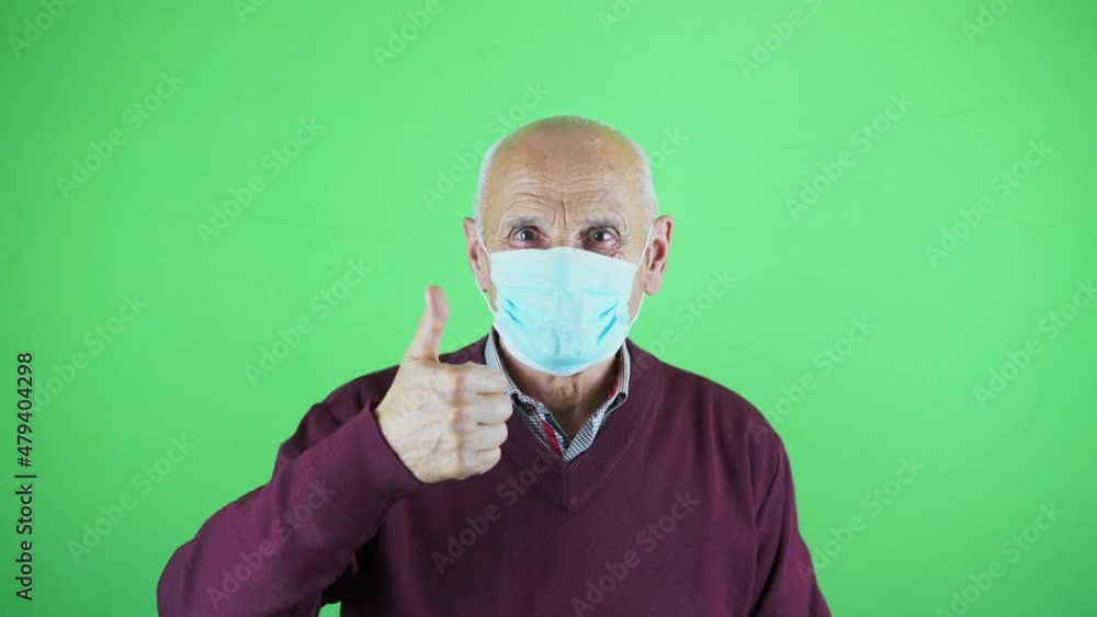 Senior man wearing face mask showing thumb up and crosses his arms over his chest on green background in studio Senior man wearing face mask showing thumb up and crosses his arms over his chest on green background in studio