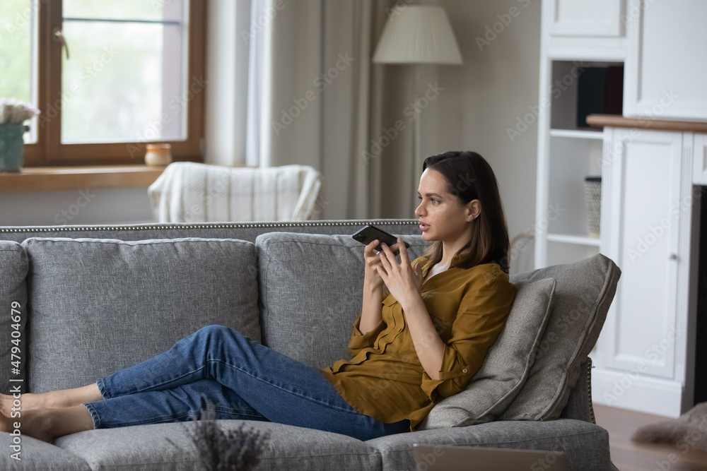 Focused millennial professional woman having mobile phone call on speaker, recording audio message for online chat on cellphone resting on couch. Business woman using voice recognition app on cell Focused millennial professional woman having mobile phone call on speaker, recording audio message for online chat on cellphone resting on couch. Business woman using voice recognition app on cell