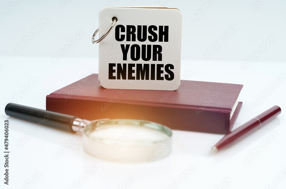 On a white surface lies a red notebook, a pen, a magnifying glass and a notebook with the inscription Crush Your Enemies