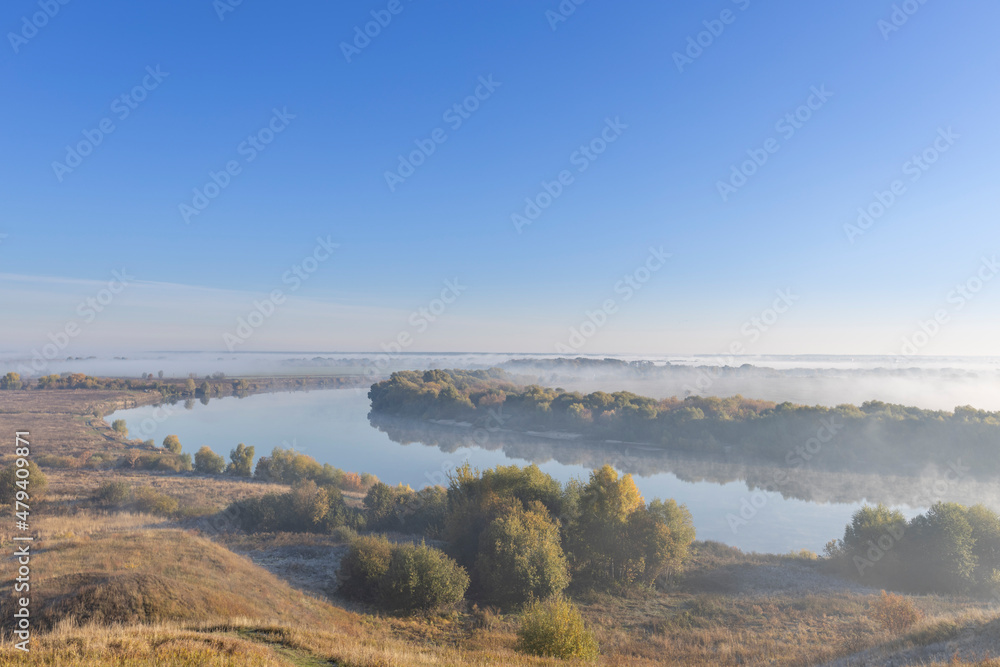 Autumn landscape in the early morning overlooking the river. A wide river and endless expanses of fields. Yellow leaves on trees and bushes are illuminated by the rays of the rising sun.