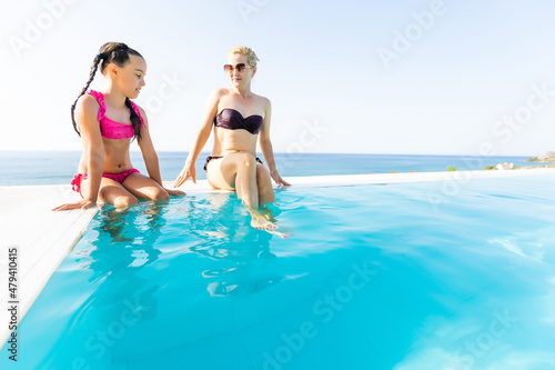 Young beautiful mother and daughter in bikini having fun resting near the pool. Happy family on summer vacation. Mom with baby girl sitting on the edge of the pool. Adventure, resort and rest concept.