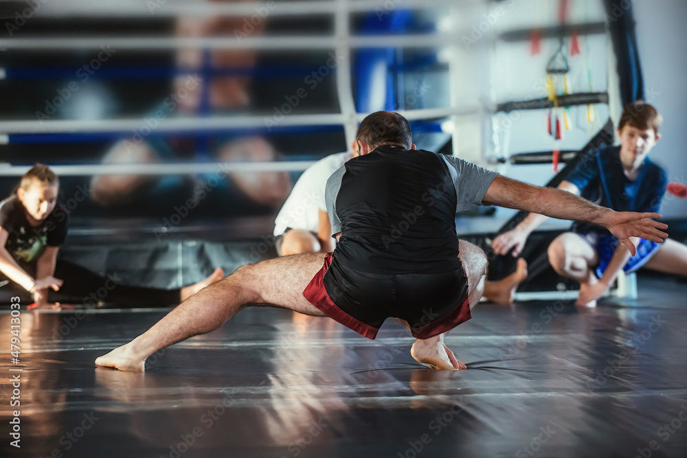 Naklejka premium The trainer conducts a training session with a group of teenage boxers. Children repeat stretching exercises after the trainer in the background of the ring.