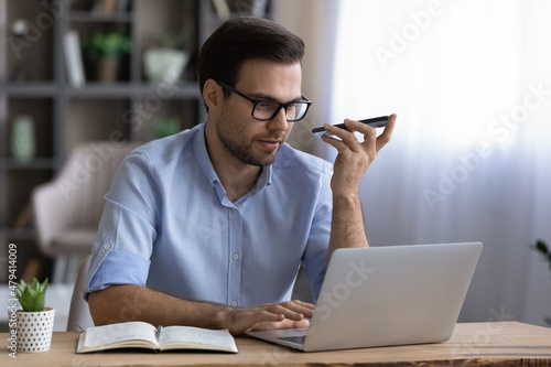 Confident focused businessman in glasses recording voice audio message on smartphone, using laptop, sitting at work desk, serious man chatting by speakerphone, activating digital assistant on device