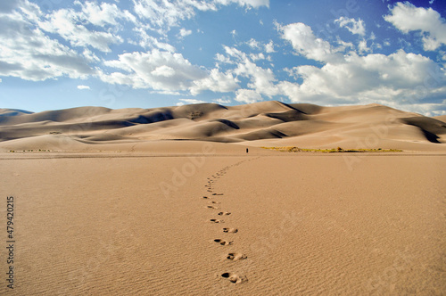 The small figure of a man walks in the distance across a desert towards towering sand dunes leaving a trail of distinct footprints behind - Great Sand Dunes National Park, Colorado - USA