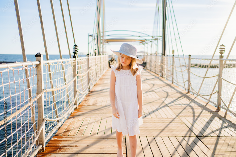 a little girl blonde in a white dress and a wide-brimmed hat on the pier
