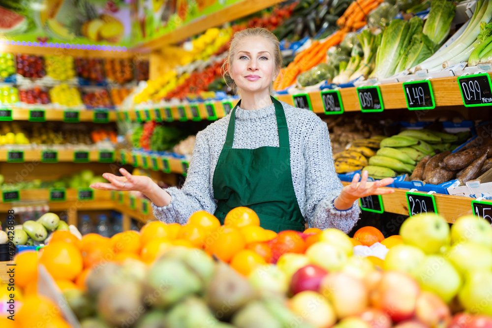 Portrait of smiling friendly saleswoman inviting to greengrocery store and offering fresh organic fruits and vegetables