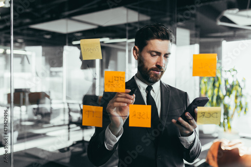 Successful businessman rewrites a task on a glass board with colored stickers, from phone and tablet