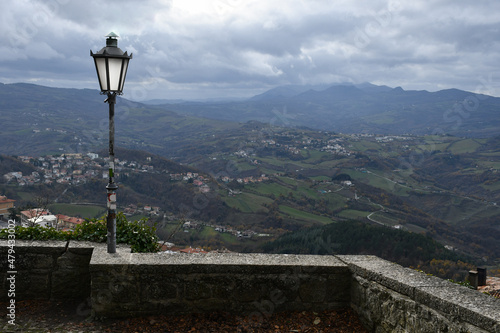 A street lamp on a stone wall against the backdrop of a mountainous landscape and houses on a cloudy day in San Marino