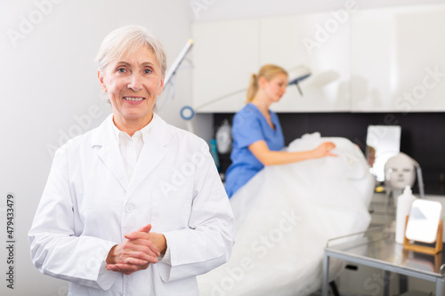 Senior woman cosmetologist standing in her office  looking at camera and smiling. Her assistant nurse working in background.