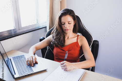 Uma mulher vestindo uma camisa vermelha sentada perto de uma janela assiste a uma aula online em seu laptop e faz anotações em seu caderno. photo