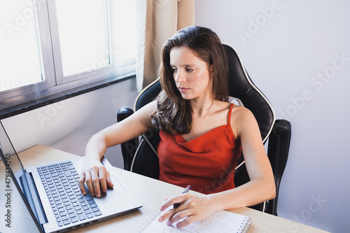 Uma mulher vestindo uma camisa vermelha sentada perto de uma janela assiste a uma aula online em seu laptop e faz anotações em seu caderno. photo