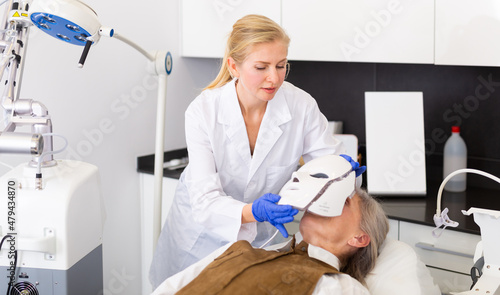 Female cosmetologist putting spectrum mask on elderly woman for photodynamic therapy. photo