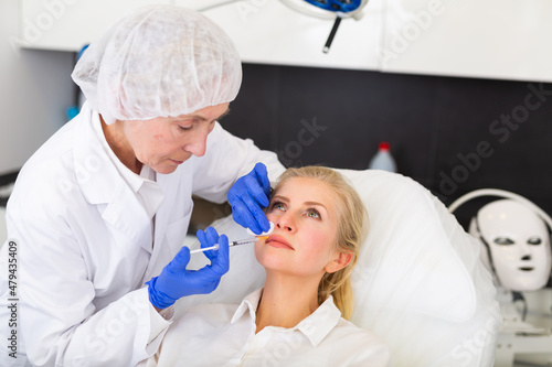 Professional experienced female cosmetologist performing face contouring injections to adult woman in aesthetic medicine clinic
