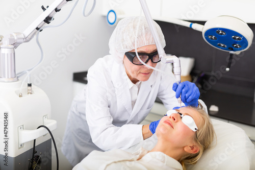 Aged woman professional beautician making procedure for laser resurfacing of female client face skin on modern equipment in clinic of aesthetic cosmetology