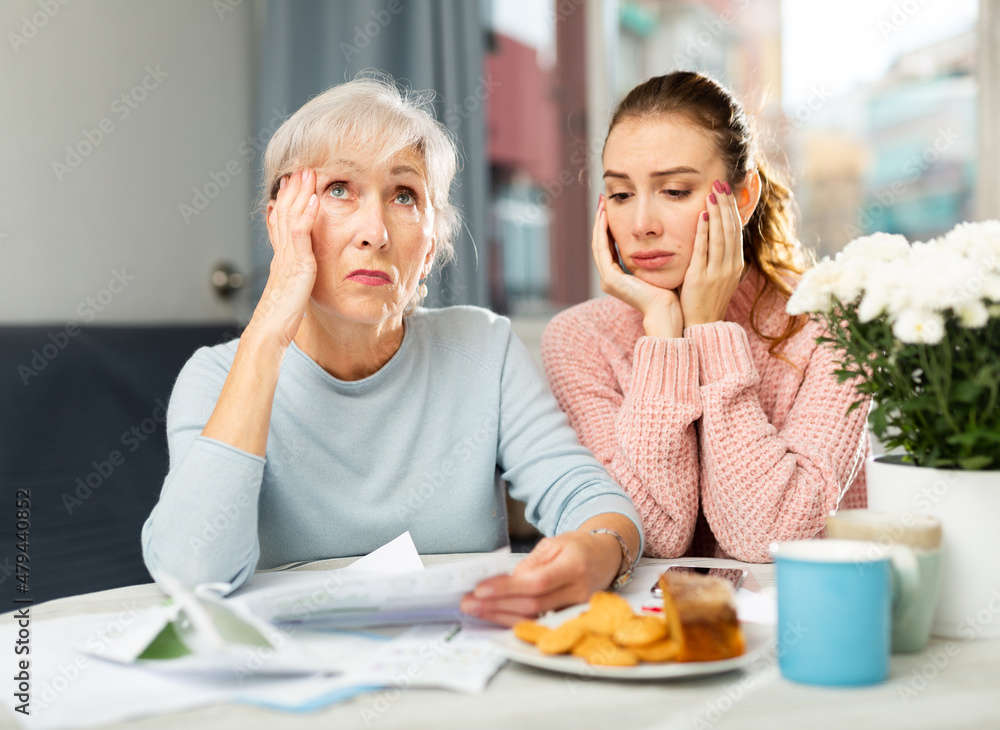 Upset young woman and her aged mother faced financials troubles, looking worriedly at papers at home.