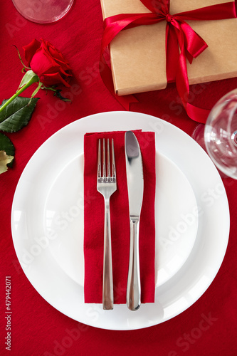 Table setting for Valentines Day romantic dinner. Top view fork and knife on red napkin on plate, rose flower, gift box, glasses on red table.