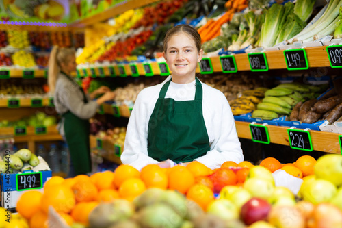 Portrait of smiling young girl wearing apron standing in fruit and vegetable store. First job concept..