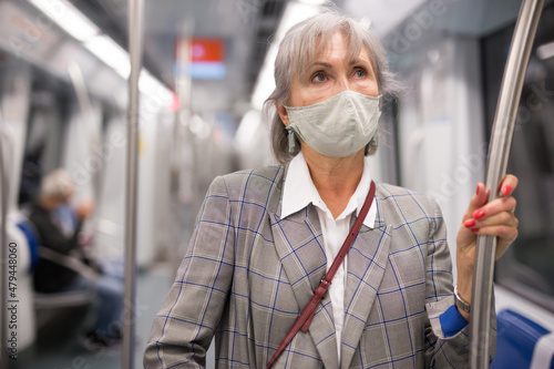 Mature European woman in face mask standing in subway train and holding handrail.