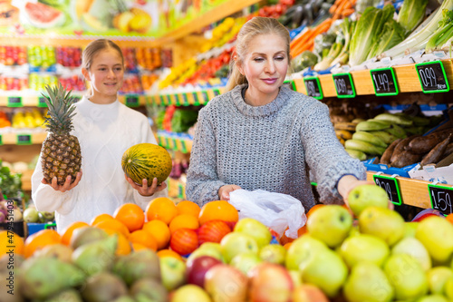 Portrait of friendly mother and teenage daughter making purchases together in greengrocery, choosing fresh fruits and vegetables