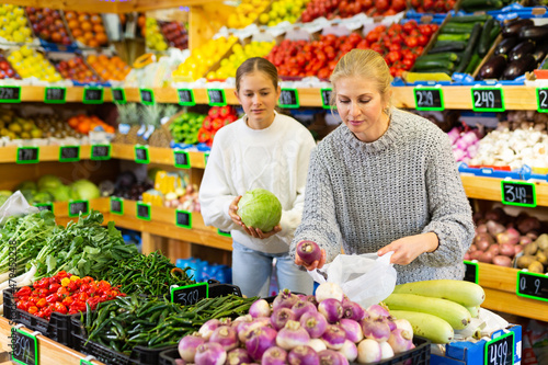 Portrait of friendly mother and teenage daughter making purchases together in greengrocery, choosing fresh fruits and vegetables