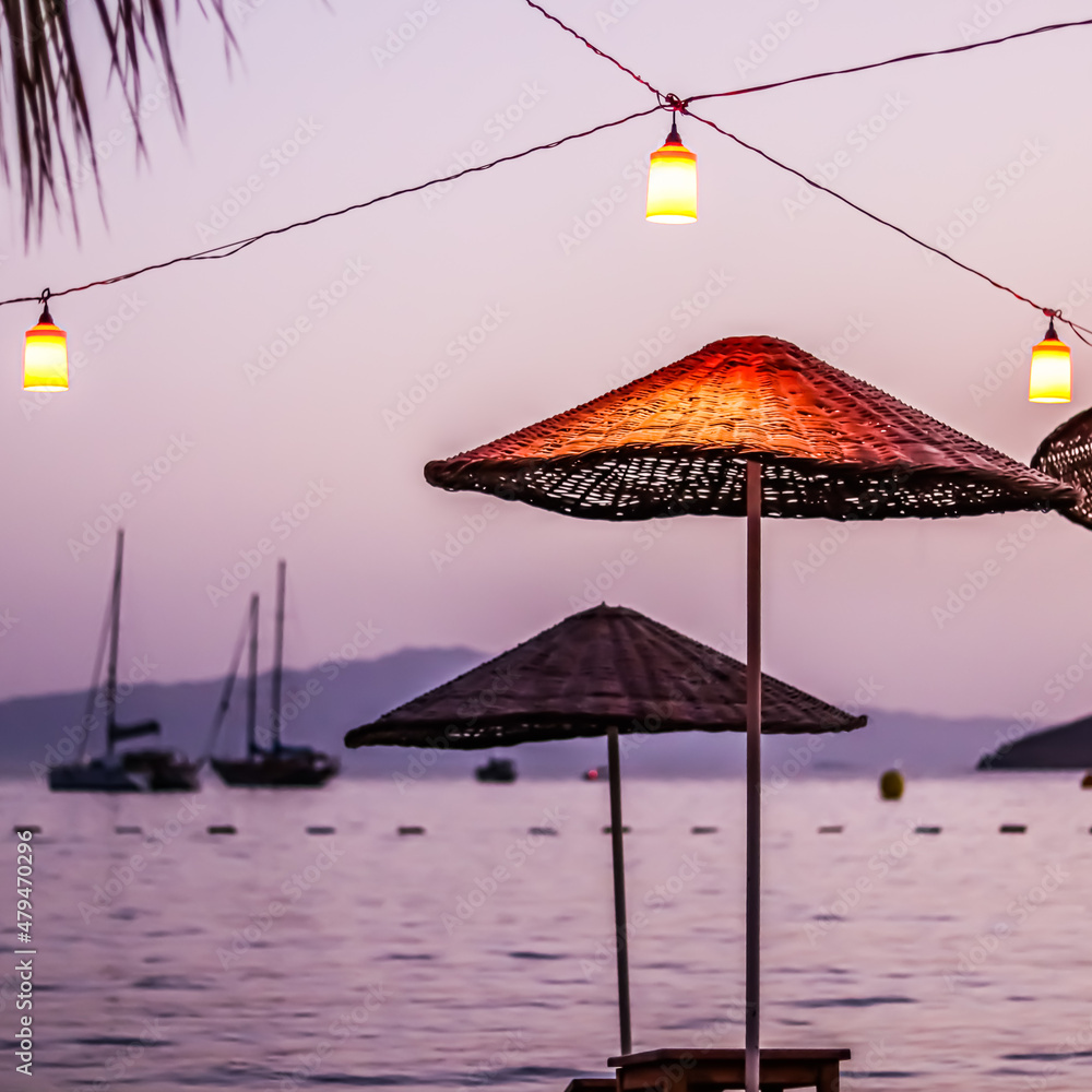 Beach umbrellas and lanterns against the backdrop of the sea, islands and yachts at sunset. Summer vacation concept
