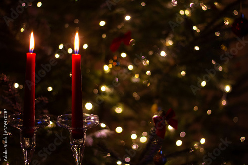 Two long red candles in tall glass candlesticks against the background of the lights of the New Year tree. A candlelit celebration in a romantic setting. Bokeh effect in the background