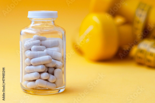Pills capsules in a glass jar close-up with dumbbells and a centimeter tape in the background. The concept of weight loss, sports, fitness, fat burner, vitamins, sports nutrition.