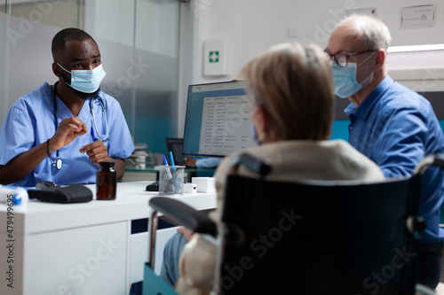 Practitioner therapist doctor with protective face mask against covid19 discussing healthcare treatment to senior woman in wheelchair. Physician man explaining sickness expertise in hospital office