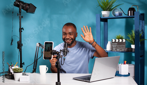 Smiling vlogger waving hello in front of recording smartphone sitting at desk with professional microphone. Content creator doing greeting hand gesture looking at live video podcast setup.