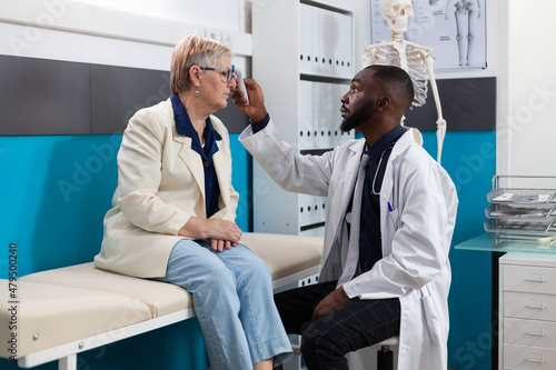 African american specialist doctor measuring temperature putting infrared thermometer on senior woman patient forehead discussing fever treatment. Medical appointment in hospital office