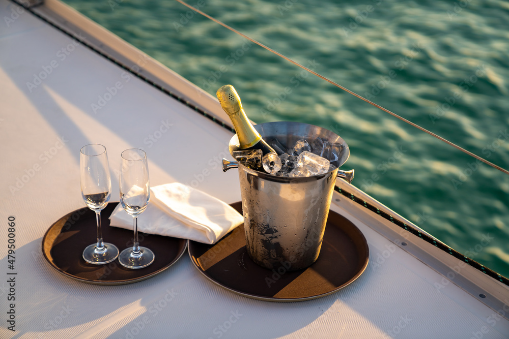 Champagne bottle in ice bucket with champagne glass for serving to passenger tourist on luxury catamaran boat sailing in the ocean at summer sunset. Tropical travel vacation sail yacht trip concept - obrazy, fototapety, plakaty 
