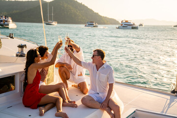 Group of man and woman friends enjoy party drinking champagne with talking together while catamaran boat sailing at summer sunset. Male and female relax outdoor lifestyle on tropical travel vacation