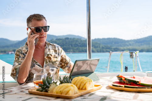 Caucasian businessman using mobile phone and working on digital tablet for online corporate business while catamaran boat sailing. Handsome man relax and enjoy outdoor lifestyle on summer vacation