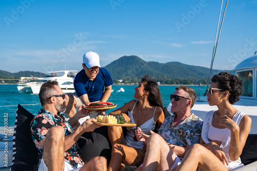 Group of Caucasian people friends enjoy luxury outdoor party eating fresh fruit together while catamaran boat sailing. Man and woman relaxing outdoor lifestyle sail yacht on summer travel vacation