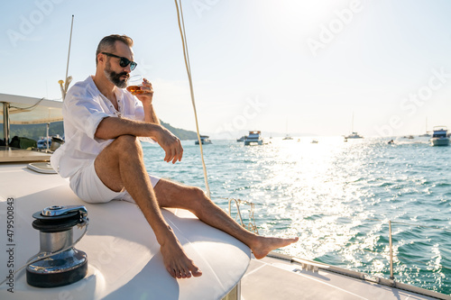 Caucasian man enjoy outdoor luxury lifestyle with alcoholic drinks while catamaran boat sailing at summer sunset. Handsome male relaxing outdoor leisure activity with tropical travel vacation trip
