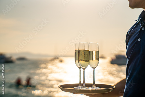 Man waiter holding champagne glass on the tray for serving to passenger tourist while luxury catamaran boat sailing in the ocean at summer sunset. Tropical travel vacation sail yacht trip concept