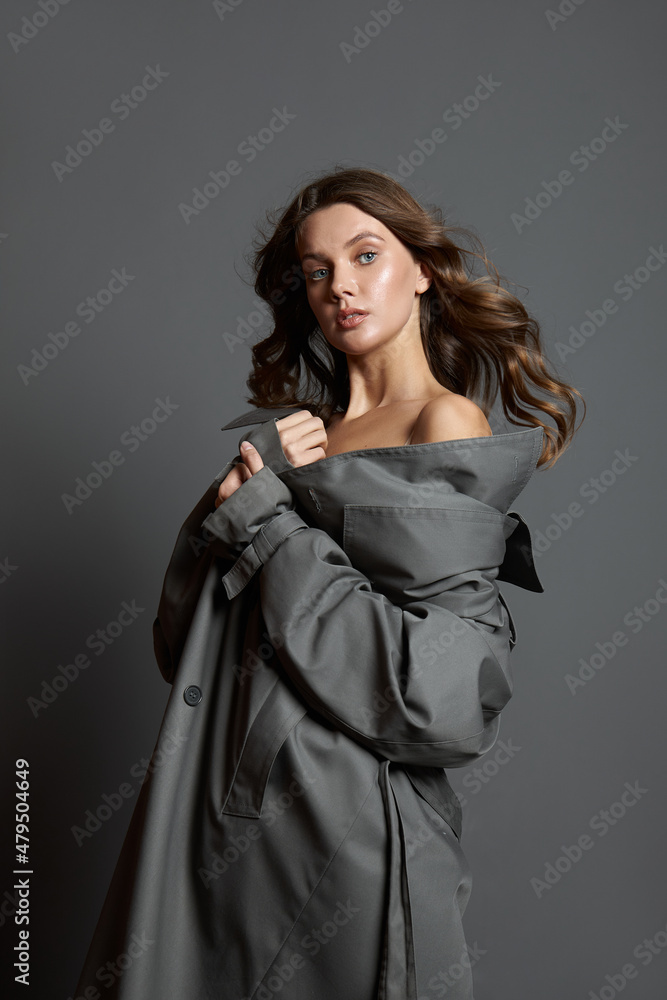 Beautiful woman in a beige bodysuit and a long gray raincoat poses standing on a gray background. Long beautiful legs Beautiful woman in a beige bodysuit and a long gray raincoat poses standing on a gray background. Long beautiful legs