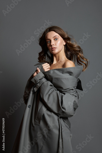 Beautiful woman in a beige bodysuit and a long gray raincoat poses standing on a gray background. Long beautiful legs