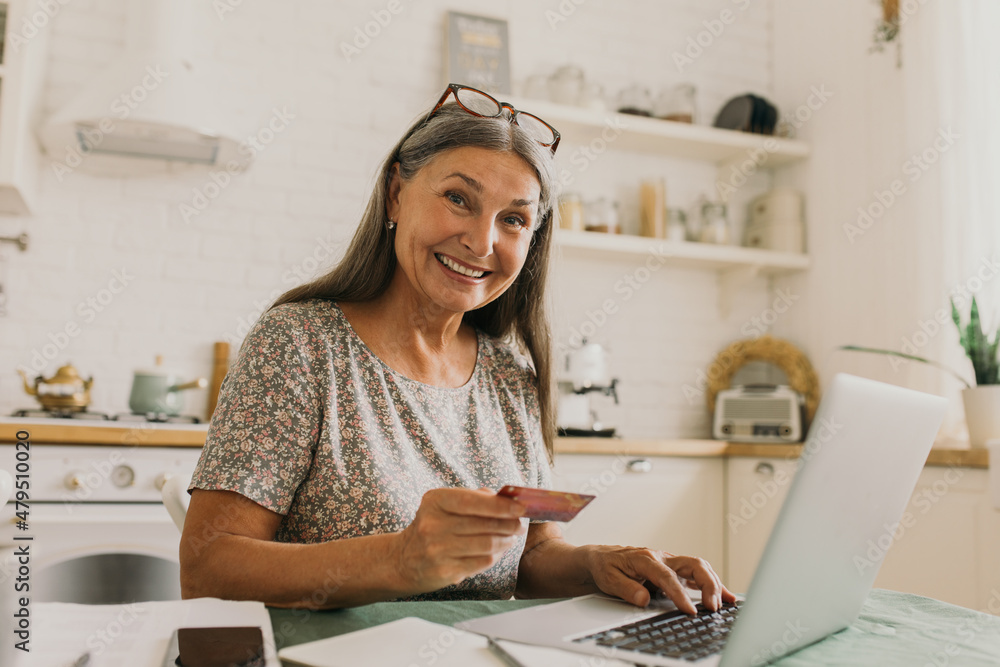 Happy senior woman paying for orders. Open laptop with information. Smiling lady sitting on kitchen, cozy home interior. Modern technology, online shopping, learn new skills concept Happy senior woman paying for orders. Open laptop with information. Smiling lady sitting on kitchen, cozy home interior. Modern technology, online shopping, learn new skills concept