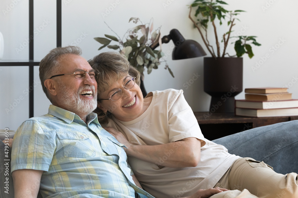Fototapeta premium Happy affectionate 60s mature family couple looking in distance, resting on comfortable sofa, enjoying communicating, dreaming planning future or watching funny TV shows together on weekend at home.