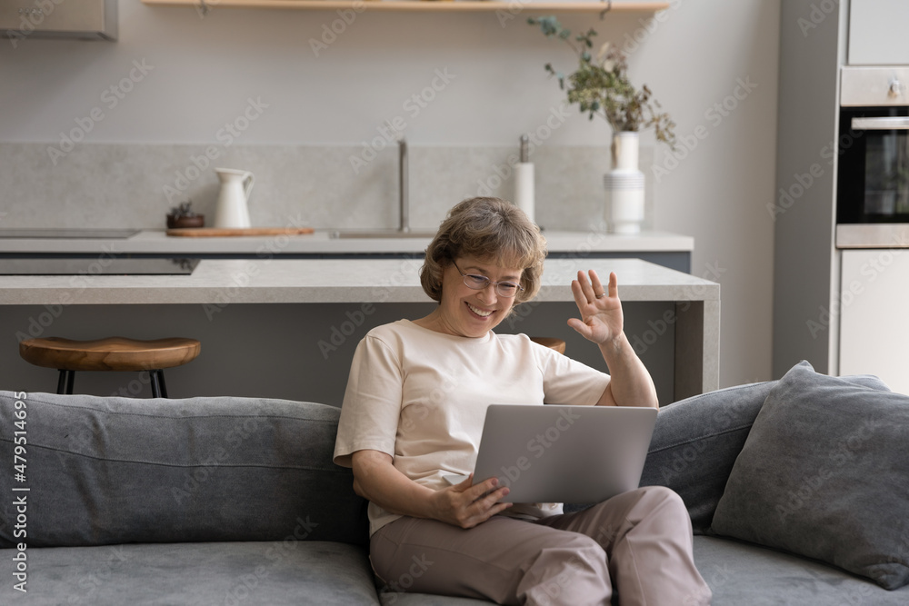 Joyful mature old retired 60s woman in eyewear waving hand, starting distant video call conversation with friends using computer app, communicating online sitting on sofa in modern living room. Joyful mature old retired 60s woman in eyewear waving hand, starting distant video call conversation with friends using computer app, communicating online sitting on sofa in modern living room.