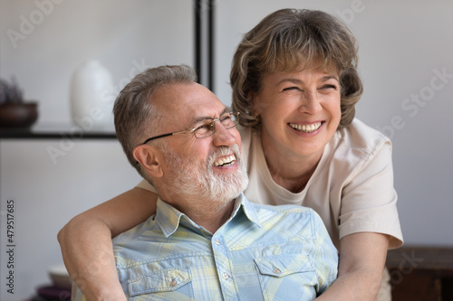 Head shot caring smiling middle aged woman cuddling happy older husband, laughing together at funny joke, looking in distance imagining future or enjoying pleasant conversation resting at home.