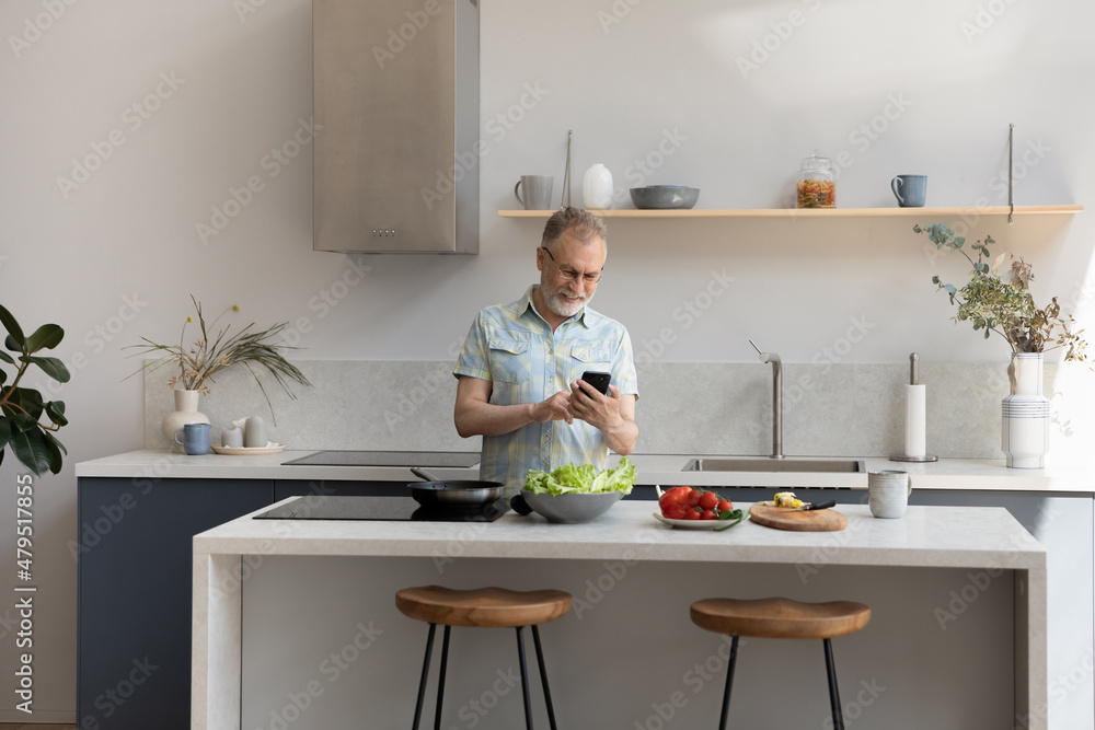 Happy middle aged 60s man distracted from preparing food using cellphone, web surfing recipe for tasty meal, communicating distantly in social network while cooking vegetarian dinner in modern kitchen Happy middle aged 60s man distracted from preparing food using cellphone, web surfing recipe for tasty meal, communicating distantly in social network while cooking vegetarian dinner in modern kitchen