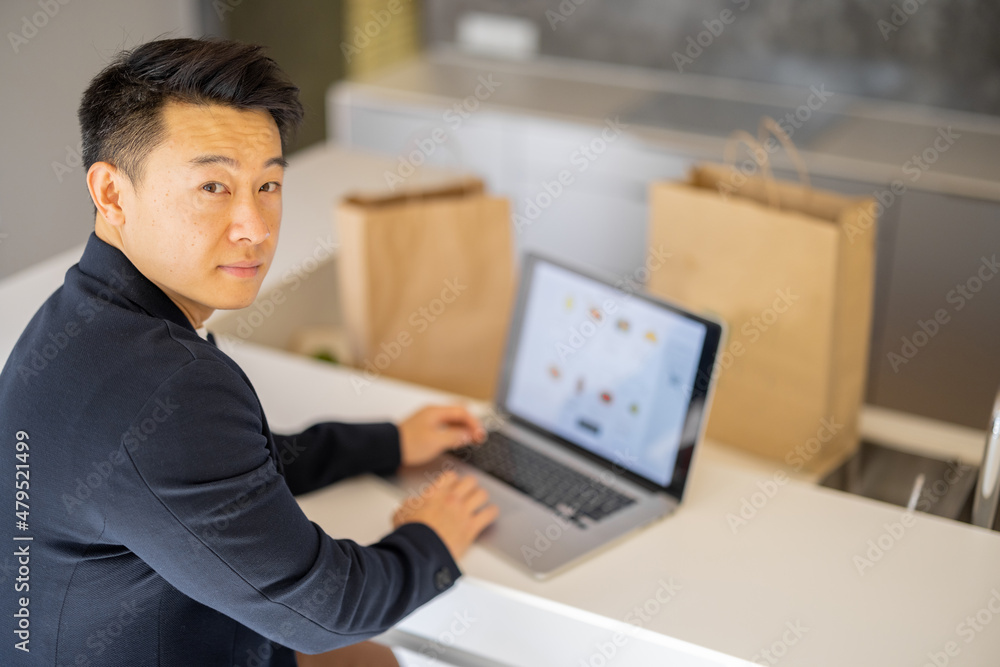 Man choosing groceries in online store on laptop computer on kitchen at home. Concept of online shopping and e-commerce. Idea of healthy and vegetarian eating. Person sit at table with shopping bags Man choosing groceries in online store on laptop computer on kitchen at home. Concept of online shopping and e-commerce. Idea of healthy and vegetarian eating. Person sit at table with shopping bags